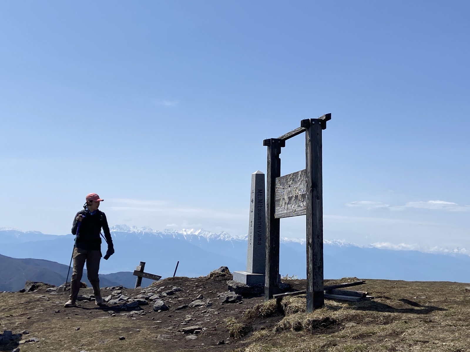 三峰山の山頂モニュメントと案内板の前を歩くピンク帽子の登山者