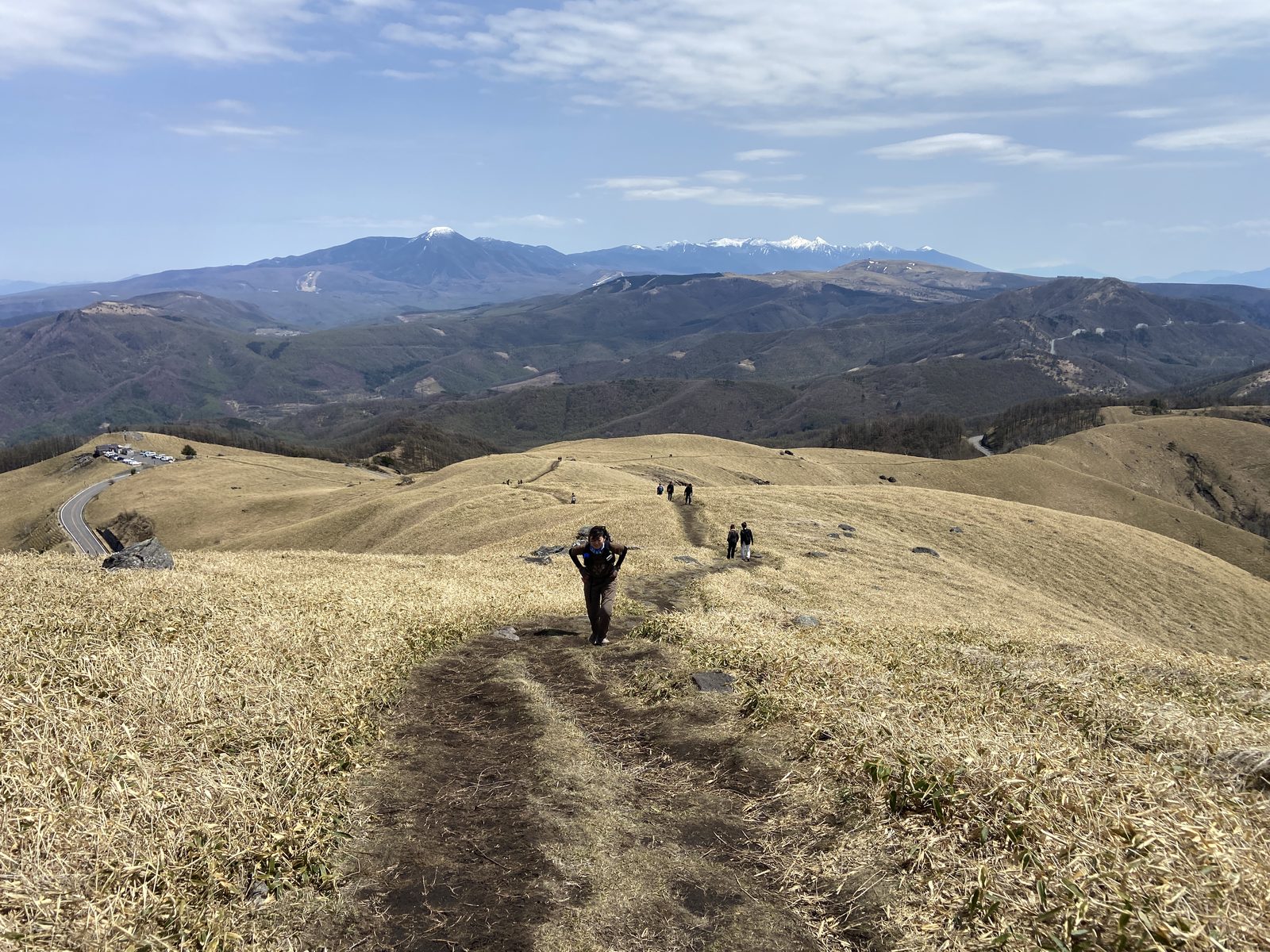 三峰山の草原の登山道を登る女性と背景の八ヶ岳と残雪の山々の信州春登山