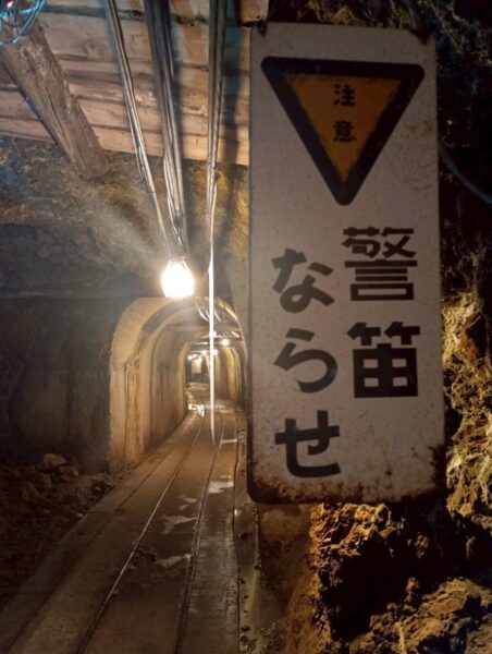 注意警笛ならせの標識とトロッコ軌道が続く佐渡金山の薄暗い坑道
