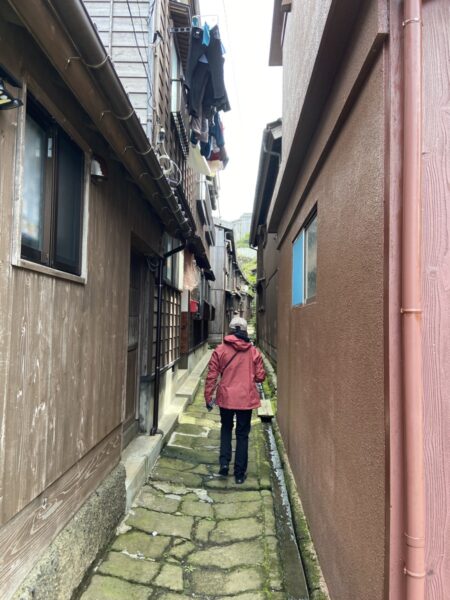 赤い上着の旅人が宿根木の石畳の路地を歩く後ろ姿、のんびり町歩き