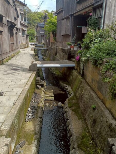 宿根木の細い水路に架かる石の橋と花、伝統的建造物が並ぶ路地