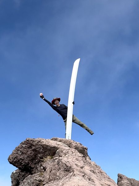 58歳の誕生日に百名山の日光白根山の山頂岩場で青空の下ポーズを決めるもおすけ、赤いハットと登山ウェアで笑顔の秋山登山
